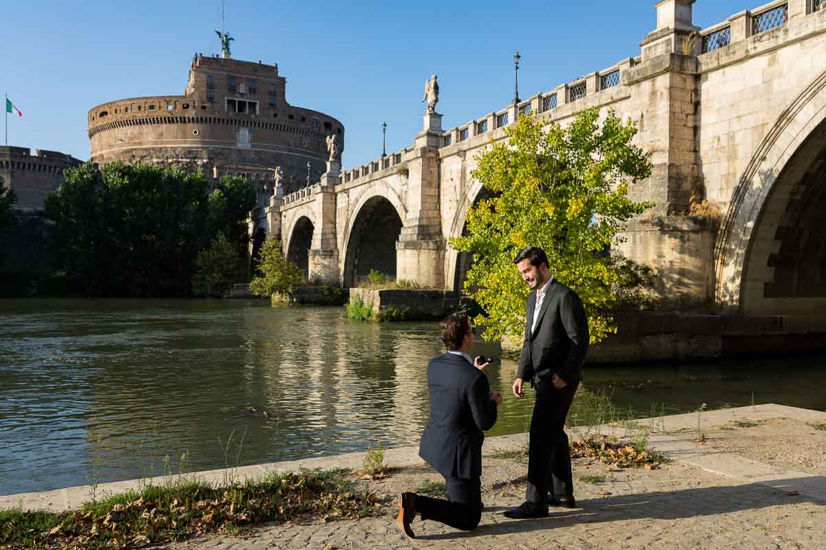 Knee down Same Sex Wedding Marriage Proposal in Rome candidly photographed on the Tiber river bank with the unique view of the Castel Sant'Angelo castle in the background