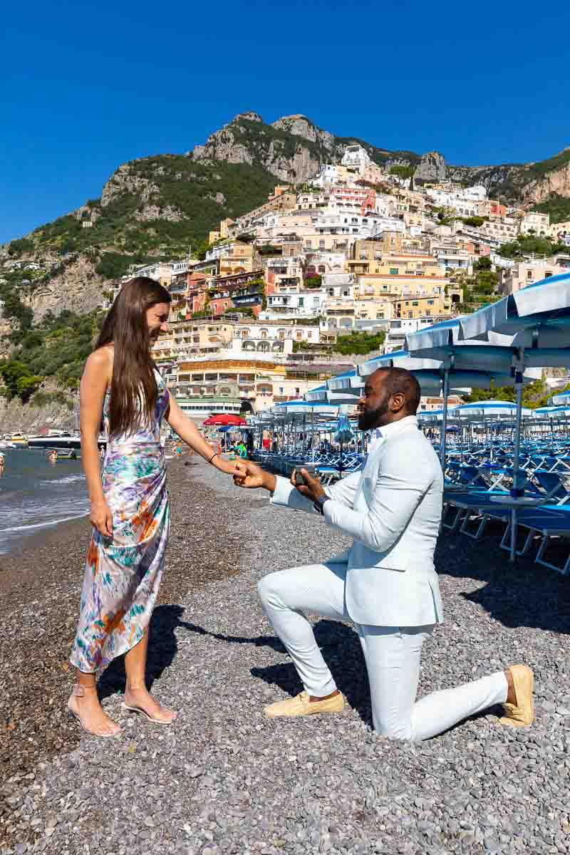 Proposing in Positano. Knee down surprise wedding proposal right beach near beach umbrellas and with the vertical town background 