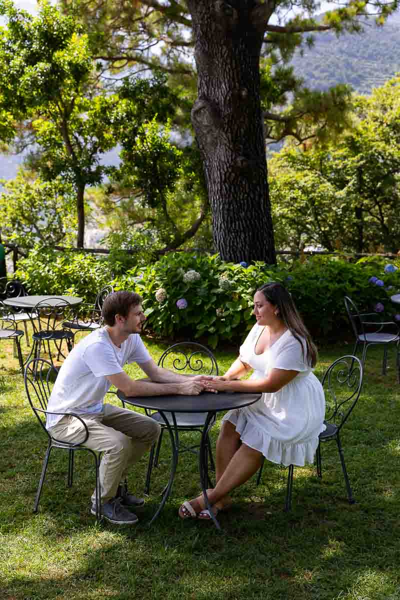 Couple sitting down holding hands in a green park with chairs chairs and trees. Posed portrait session