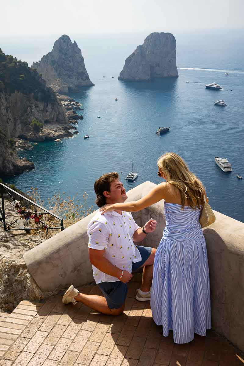 Kneeling down for a marriage engagement overlooking the Faraglioni rocks far distance 