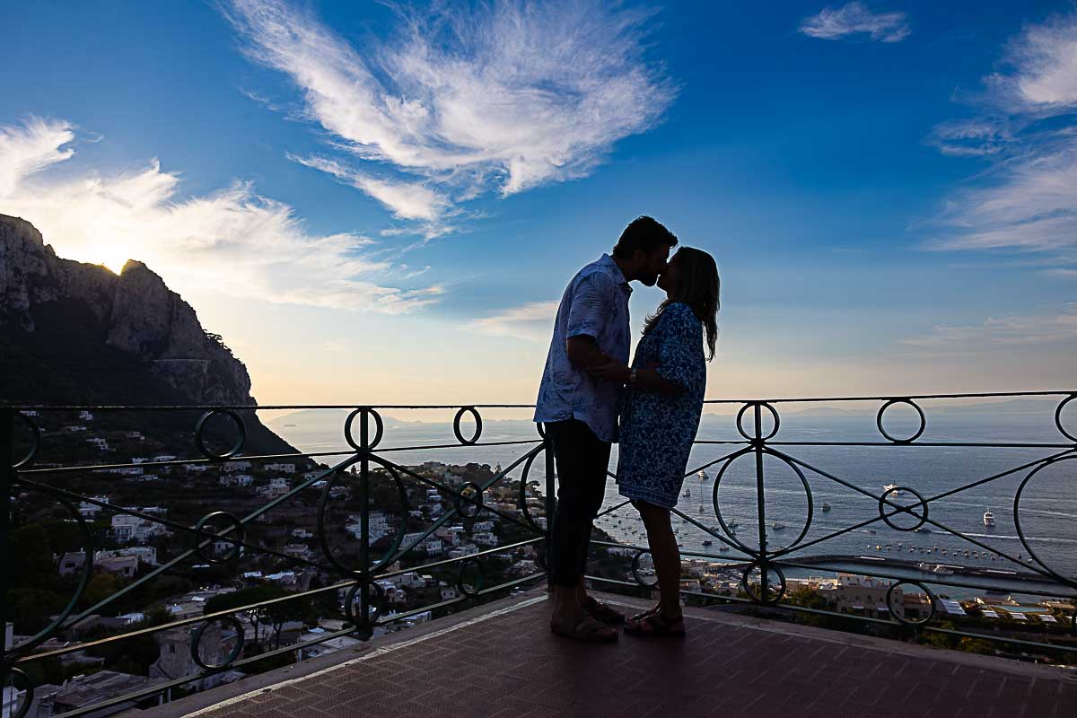 In love kissing while taking pictures on a terrace overlooking the sea at sunset with a photographer taking photos 