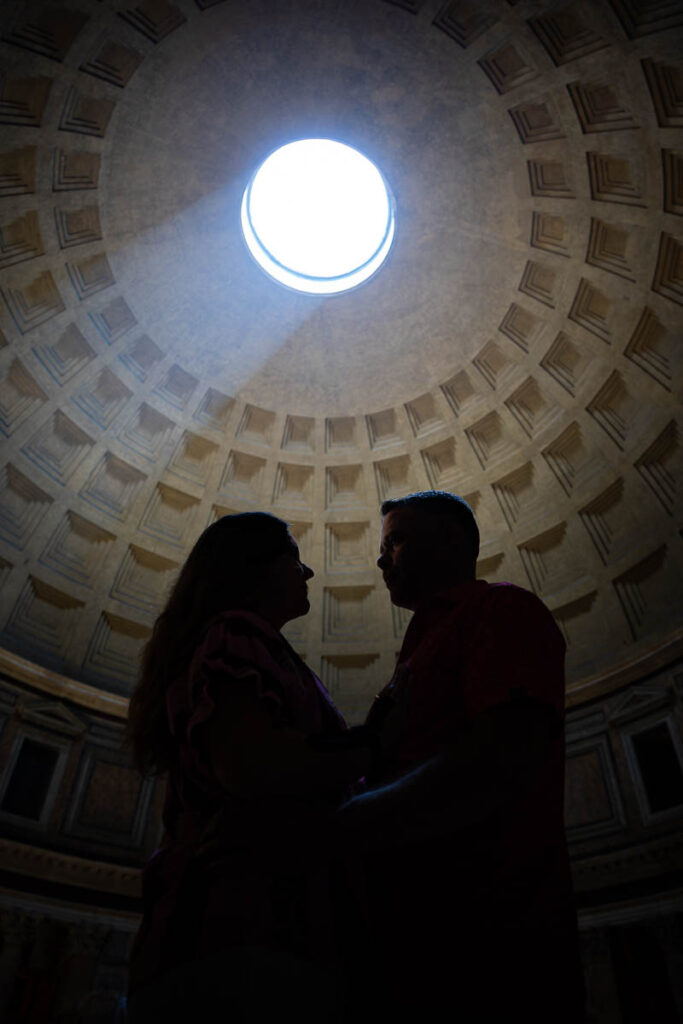 Silhouette of an engaged couple standing close beneath the dome with dramatic backlighting from the sunbeam above, captured in an artistic architectural photography composition