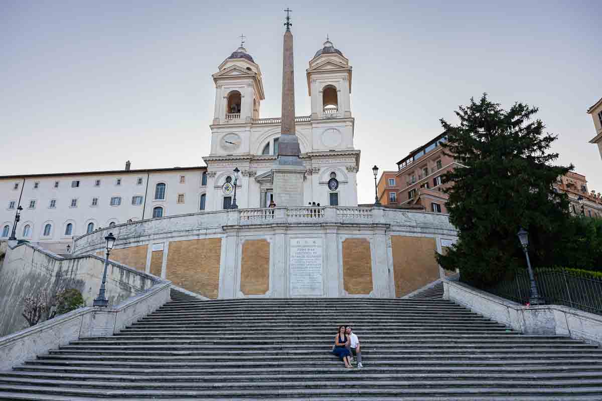 Couple seated high along Spanish Steps beneath Trinità dei Monti church, framed as small figures within a wide staircase scene creating a cinematic composition with strong perspective, scale, and depth.