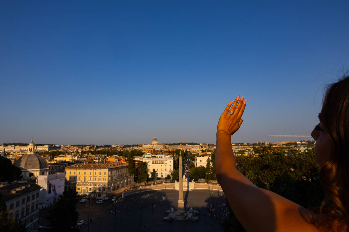 Newly engaged couple celebrating with a raised hand displaying the diamond ring above Piazza del Popolo, captured in a creative close-up composition with the cityscape of Rome creating a memorable backdrop.