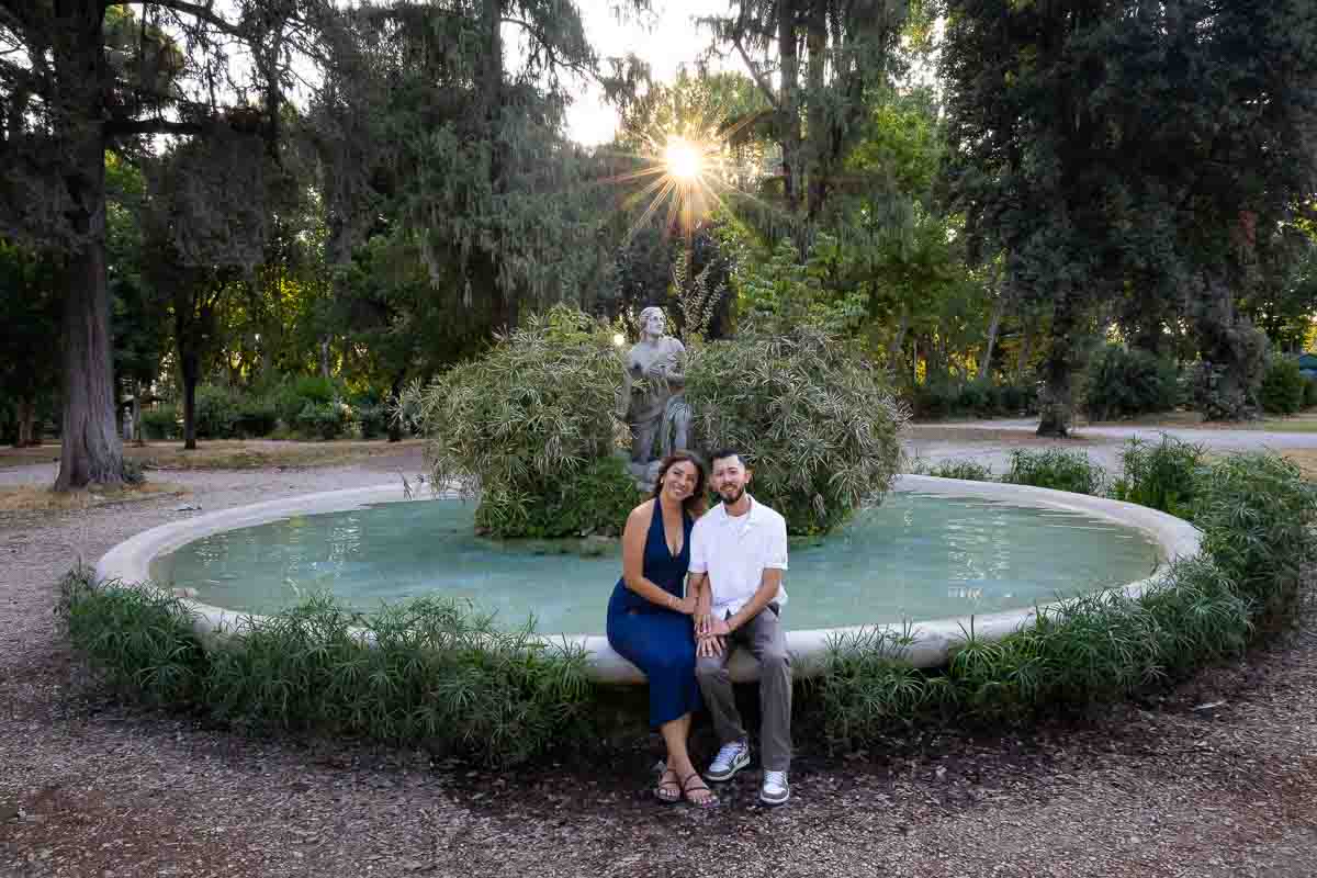 Couple seated beside a water fountain during a romantic portrait session at sunset, photographed with a narrow aperture technique to capture defined light rays, sun flare details, and a warm atmospheric composition.