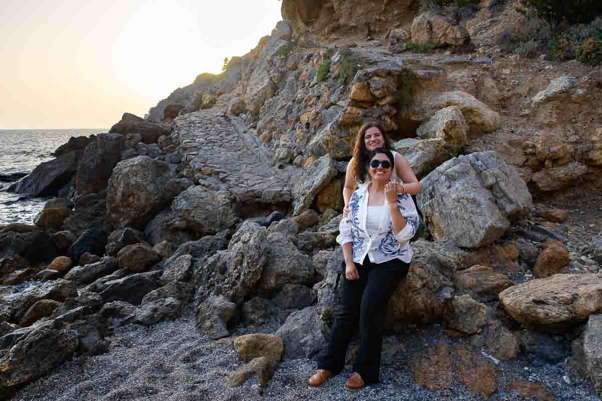 Golden hour portrait of two women leaning against a large coastal boulder during an engagement photography session.