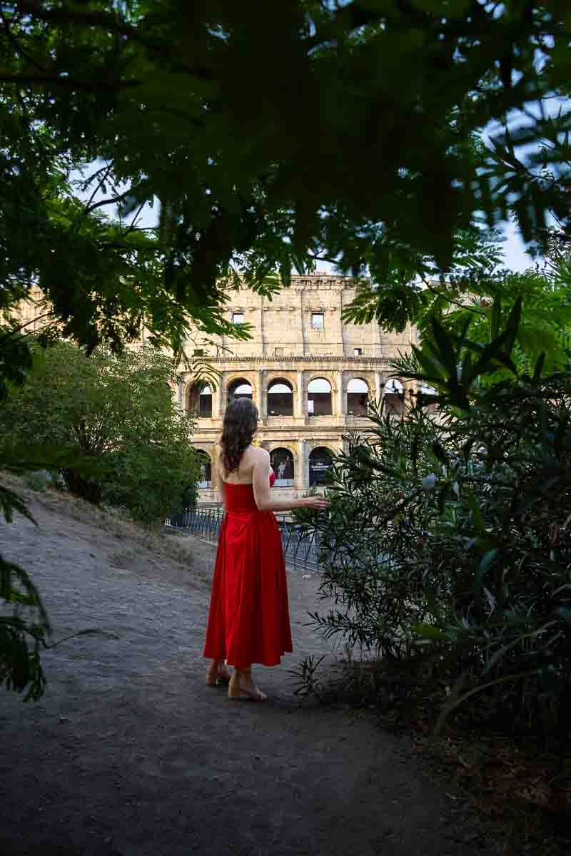 Natural travel portrait of a woman standing among garden greenery while admiring the ancient amphitheatre from a quiet elevated viewpoint, photographed with a relaxed outdoor composition.